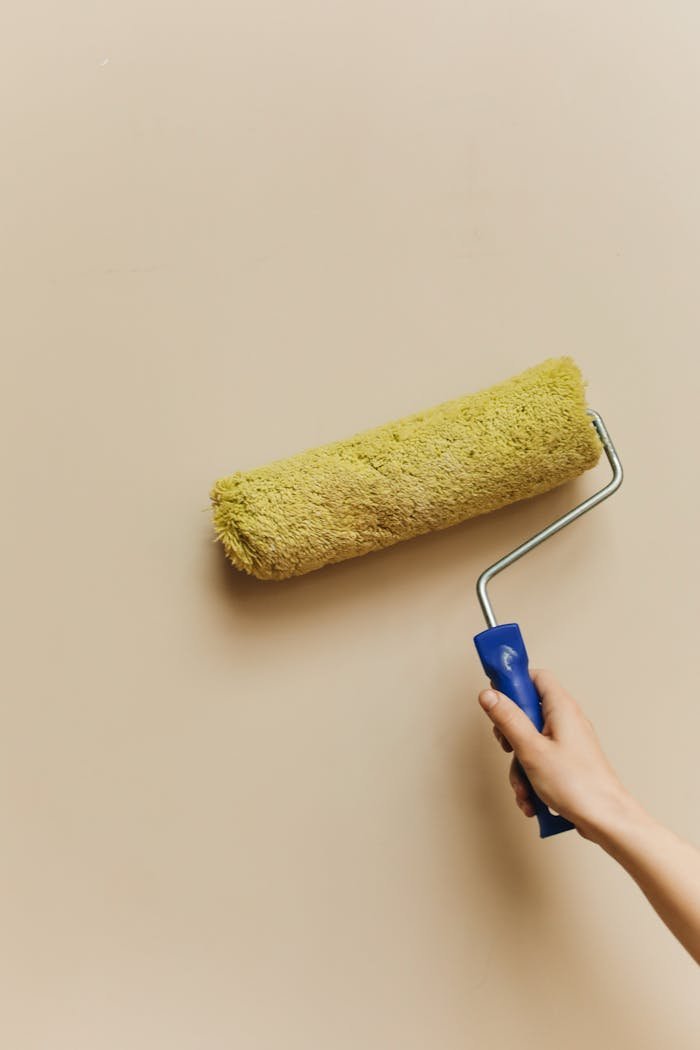 Hand holding a paint roller applying fresh coat on beige wall in home improvement project.
